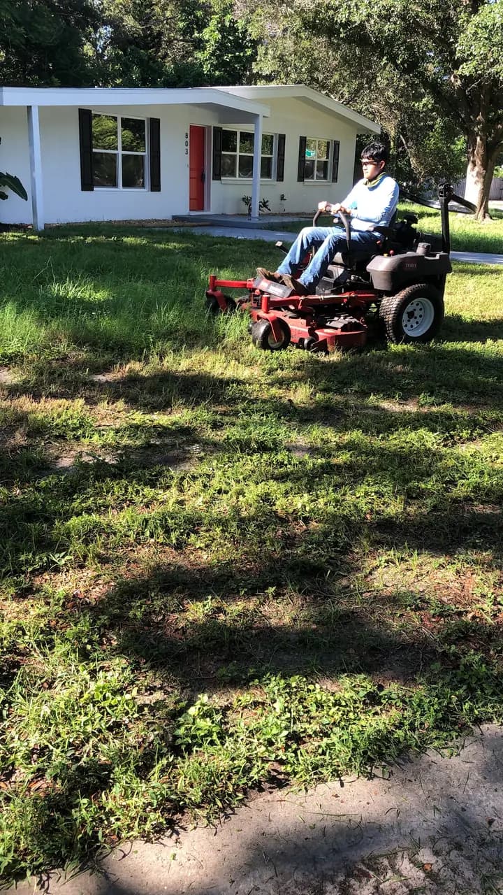 Crew member on a zero-turn mower in front of a white ranch home